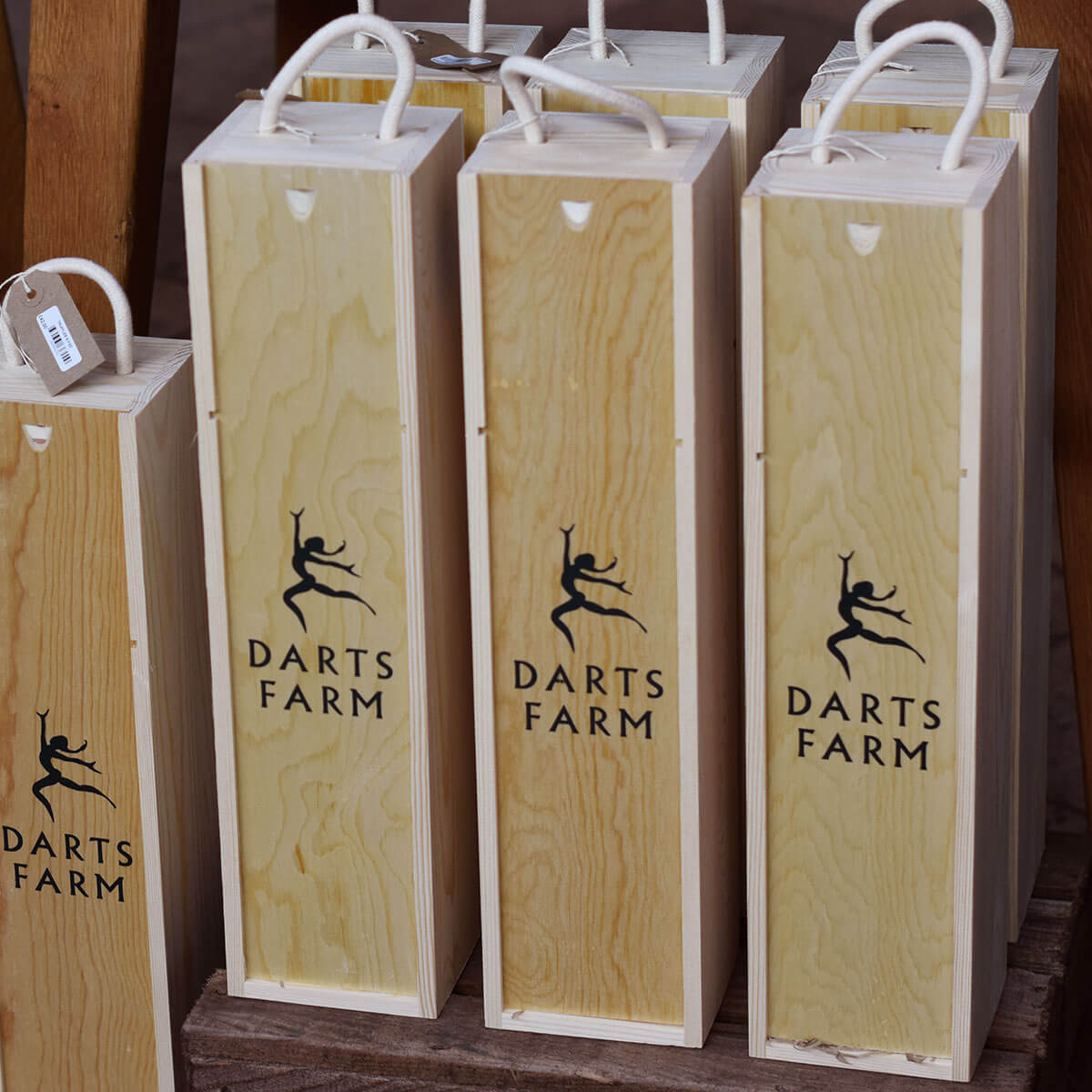 Several tall, narrow wooden darts boxes with white paper handles are stacked on a wooden surface. The front boxes are light natural wood with a black printed Darts Farm logo and text, and a small product tag is attached to one box.