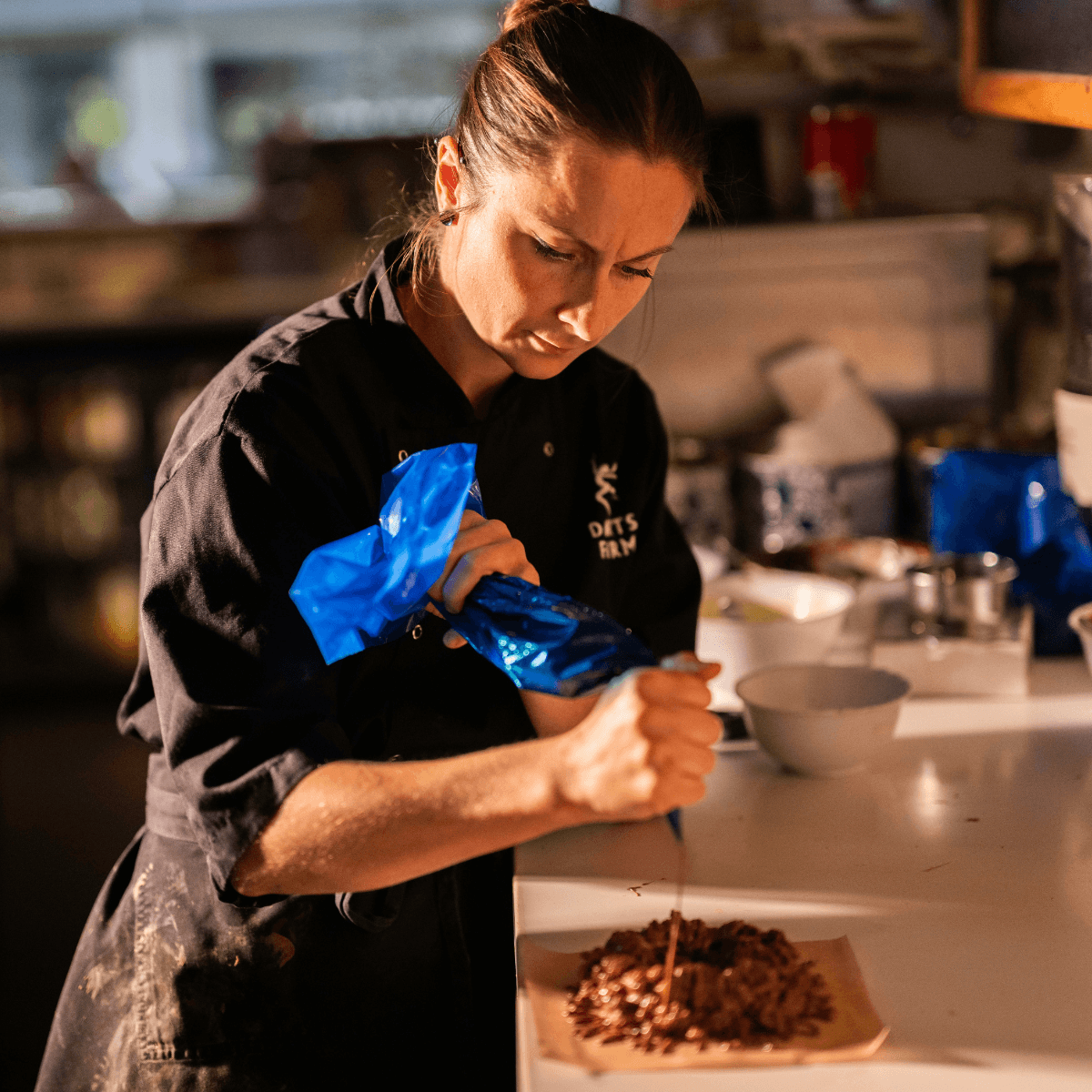 A chef in a black uniform pipes chocolate onto a plate in a warm, softly lit kitchen setting.