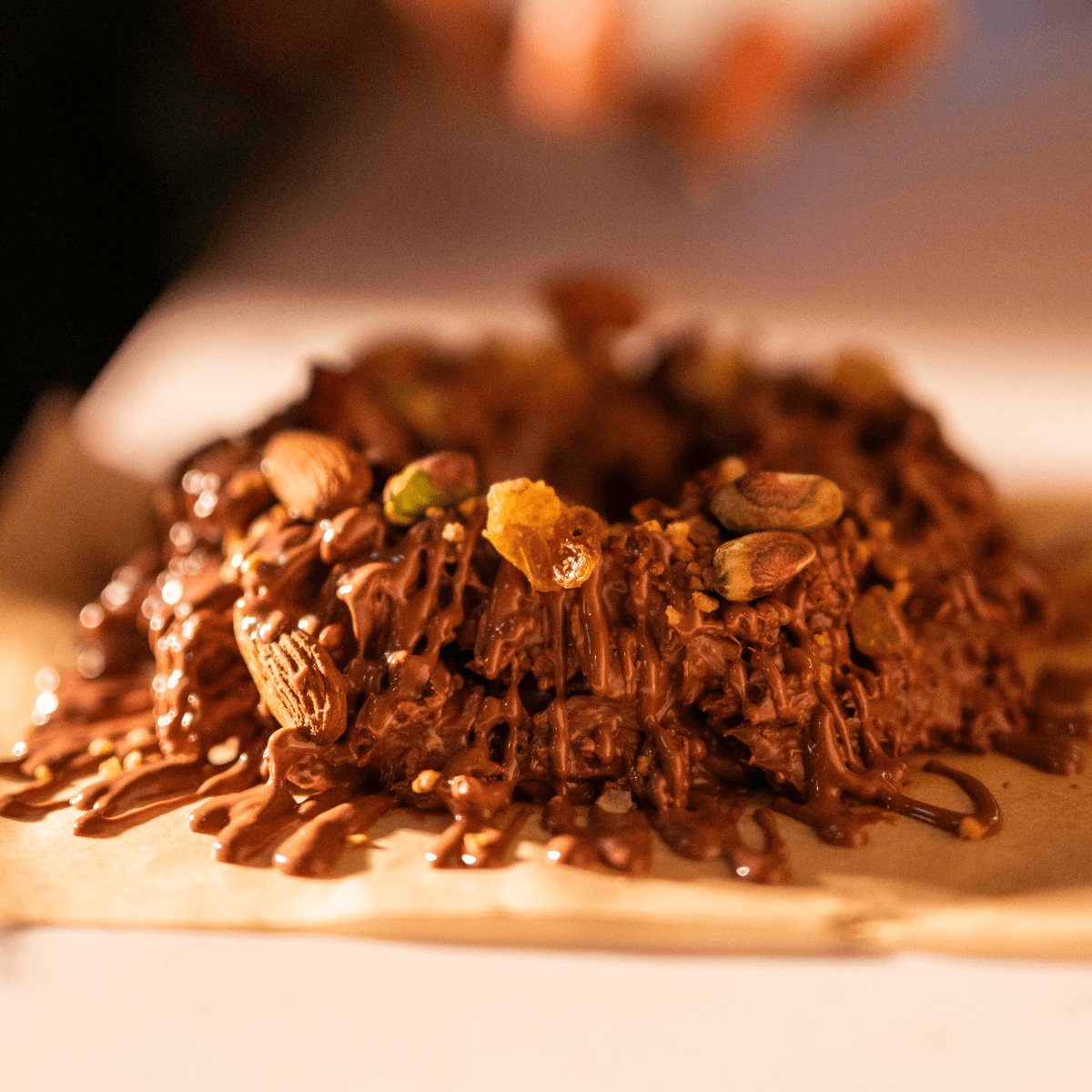 A close-up of a chocolate-covered confection topped with almonds, pistachios, and dried fruit, resting on parchment paper with a warm, softly blurred background.